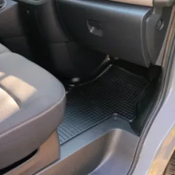 Wnętrze samochodu z szarą tapicerką foteli i czarną gumową wycieraczką na podłodze. Widoczne detale deski rozdzielczej i otwarte drzwi.