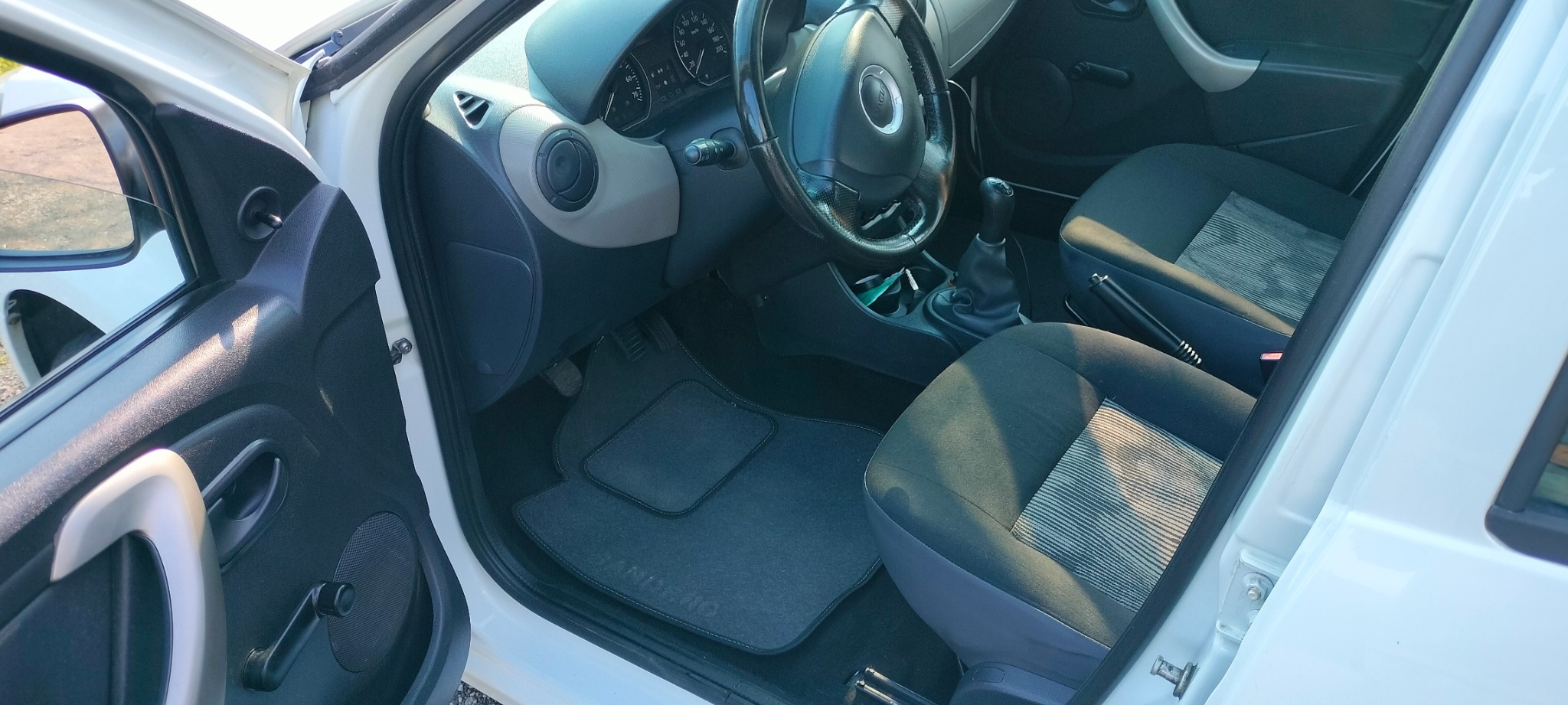 Wnętrze białego samochodu z czarną tapicerką i szarymi dywanikami. Widoczne fotele, deska rozdzielcza i kierownica. Czyste i zadbane wnętrze pojazdu.