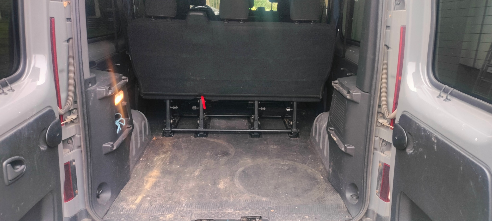 Wnętrze bagażnika szarego busa z widocznymi śladami użytkowania na podłodze. Tylne siedzenia zamontowane. Otwarte drzwi bagażnika z widocznymi elementami oświetlenia.