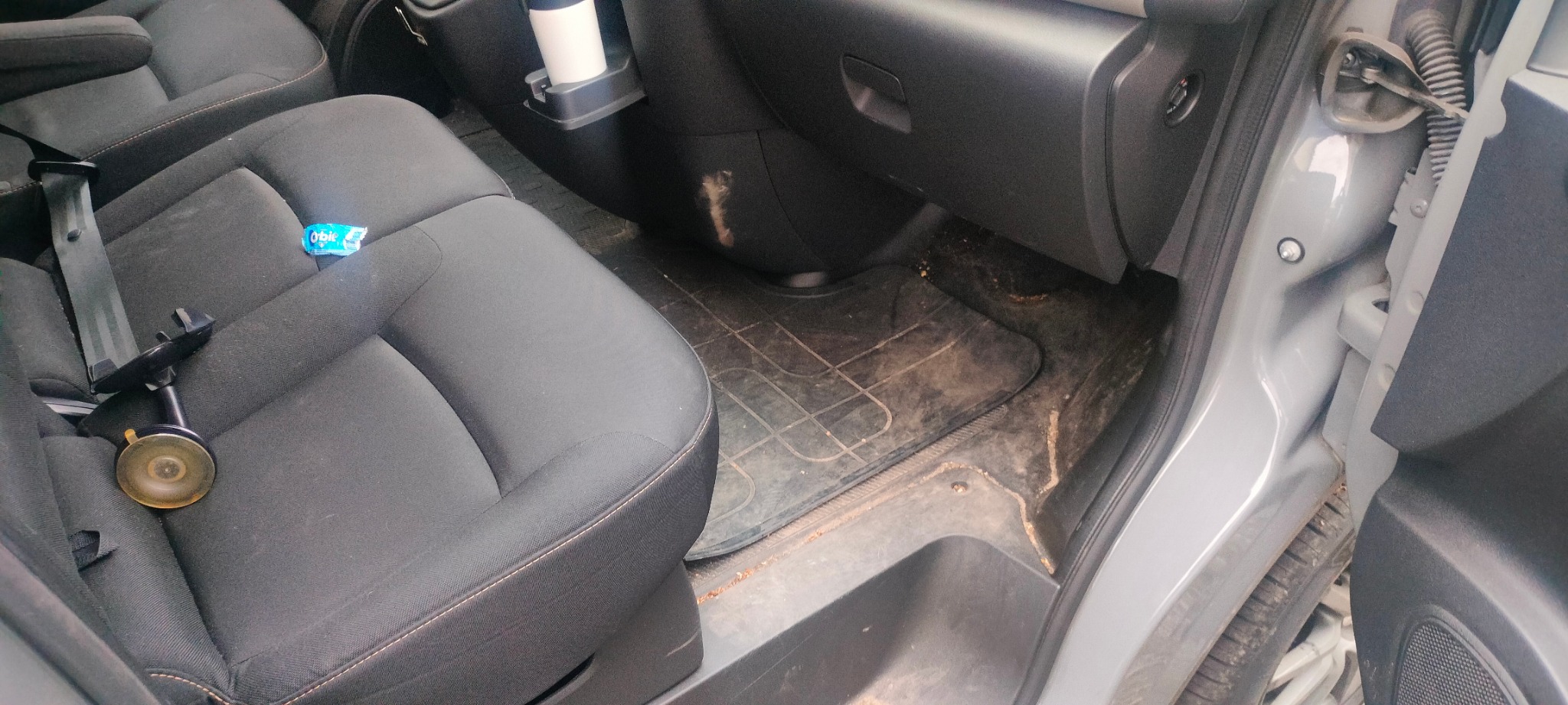 Wnętrze brudnego samochodu z czarną tapicerką, gdzie widoczny jest brud na dywaniku, siedzeniu i plastikowych elementach. Na siedzeniu leży guma do żucia.
