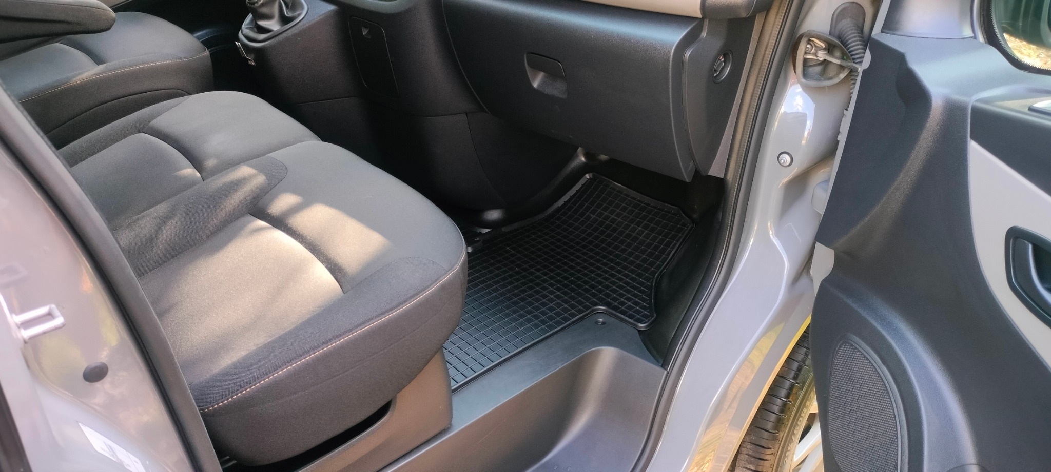 Wnętrze samochodu z szarą tapicerką foteli i czarną gumową wycieraczką na podłodze. Widoczne detale deski rozdzielczej i otwarte drzwi.
