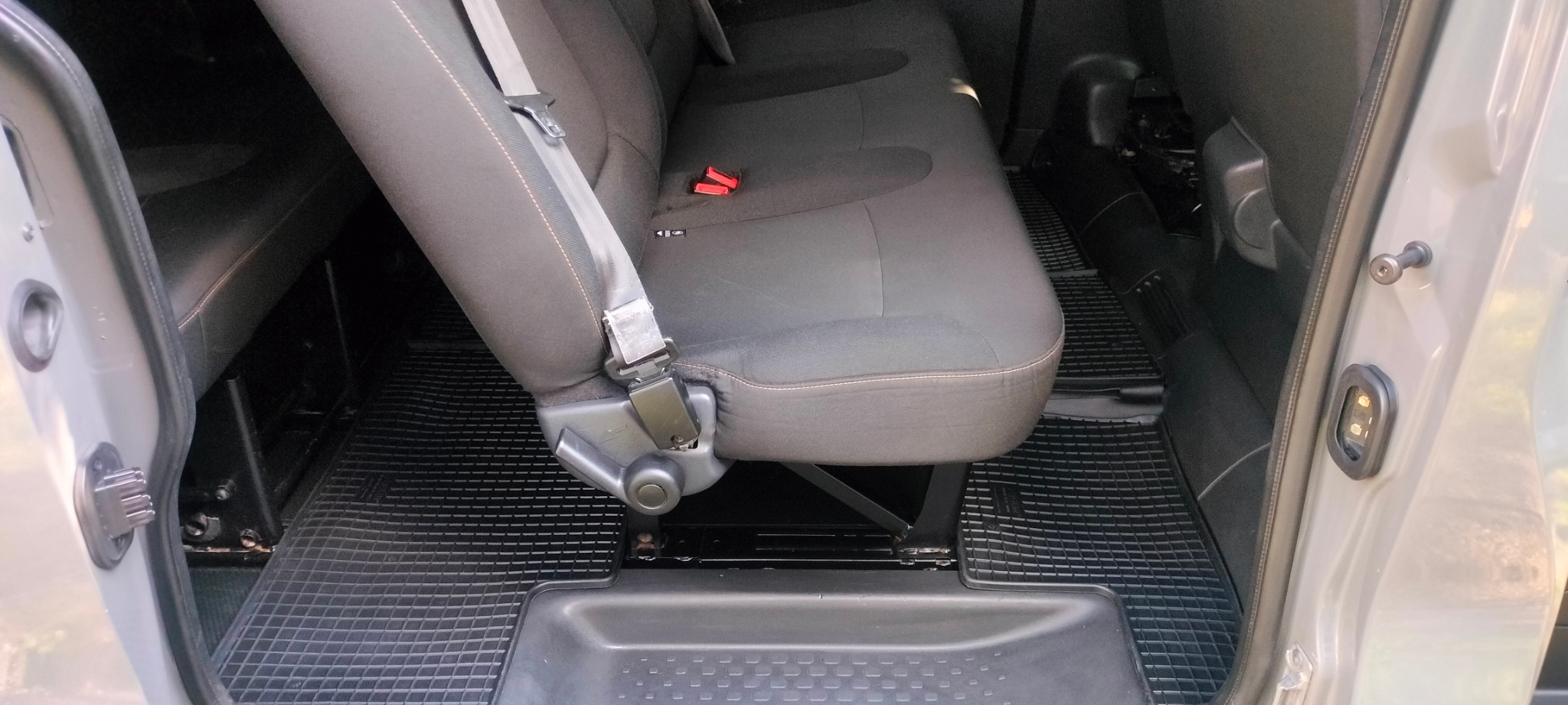 Szare, tapicerowane siedzenie w samochodzie z pasami bezpieczeństwa i gumową wycieraczką podłogową. Widok z wnętrza pojazdu przez otwarte drzwi.
