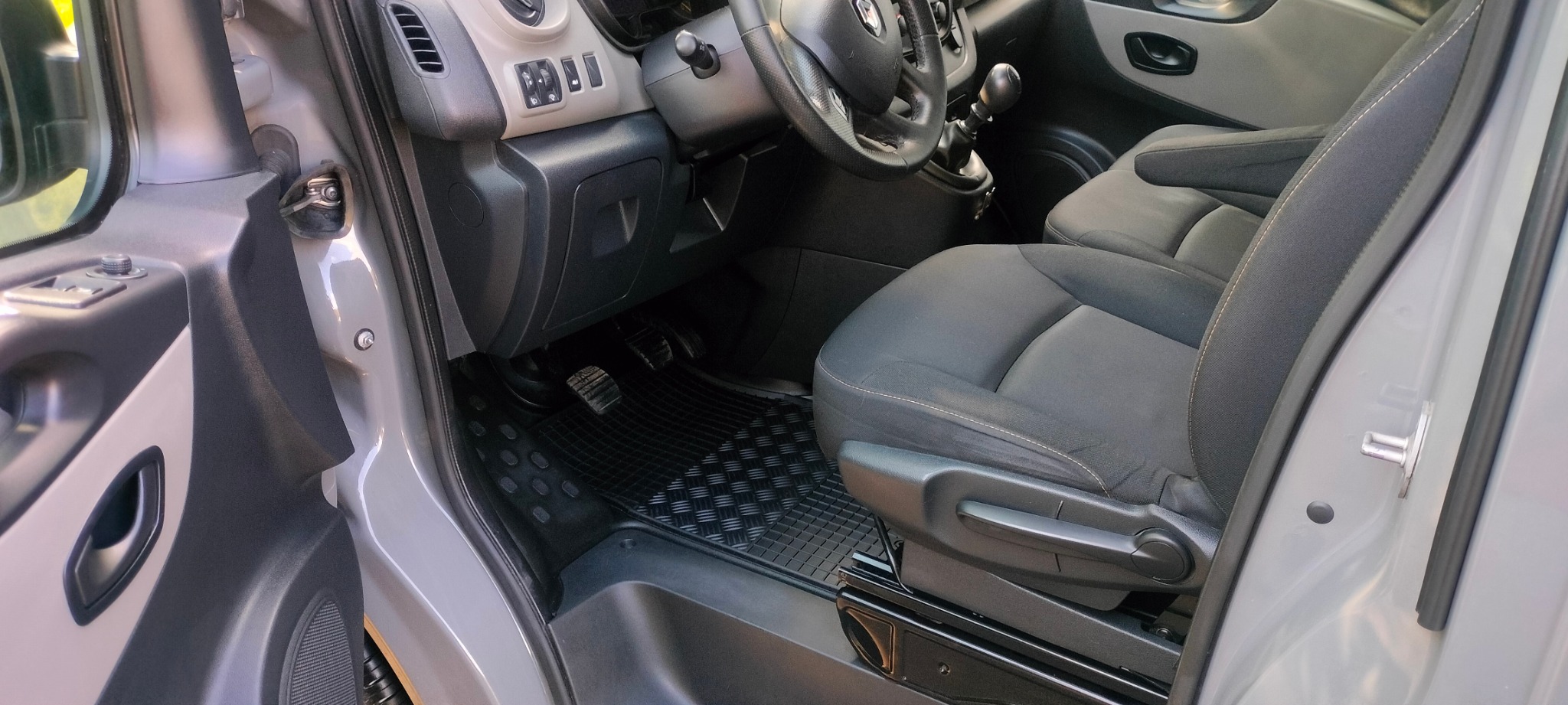 Wnętrze szarego samochodu z widocznym fotelem kierowcy, czarnymi dywanikami i deską rozdzielczą. Czyste, zadbane wnętrze pojazdu z otwartymi drzwiami.