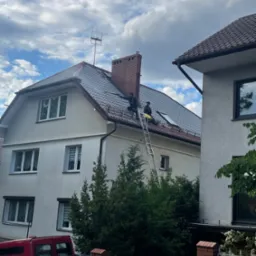 Dwóch dekarzy na drabinie przy dachu dwupiętrowego domu z ceglanym kominem i anteną. Widok na szare dachówki i okno dachowe. Na dole widoczny czerwony samochód.