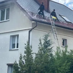 Dwóch mężczyzn czyści dach domu z metalową dachówką za pomocą myjki ciśnieniowej, stojąc na drabinie, z widocznym ceglanym kominem i oknami dachowymi.