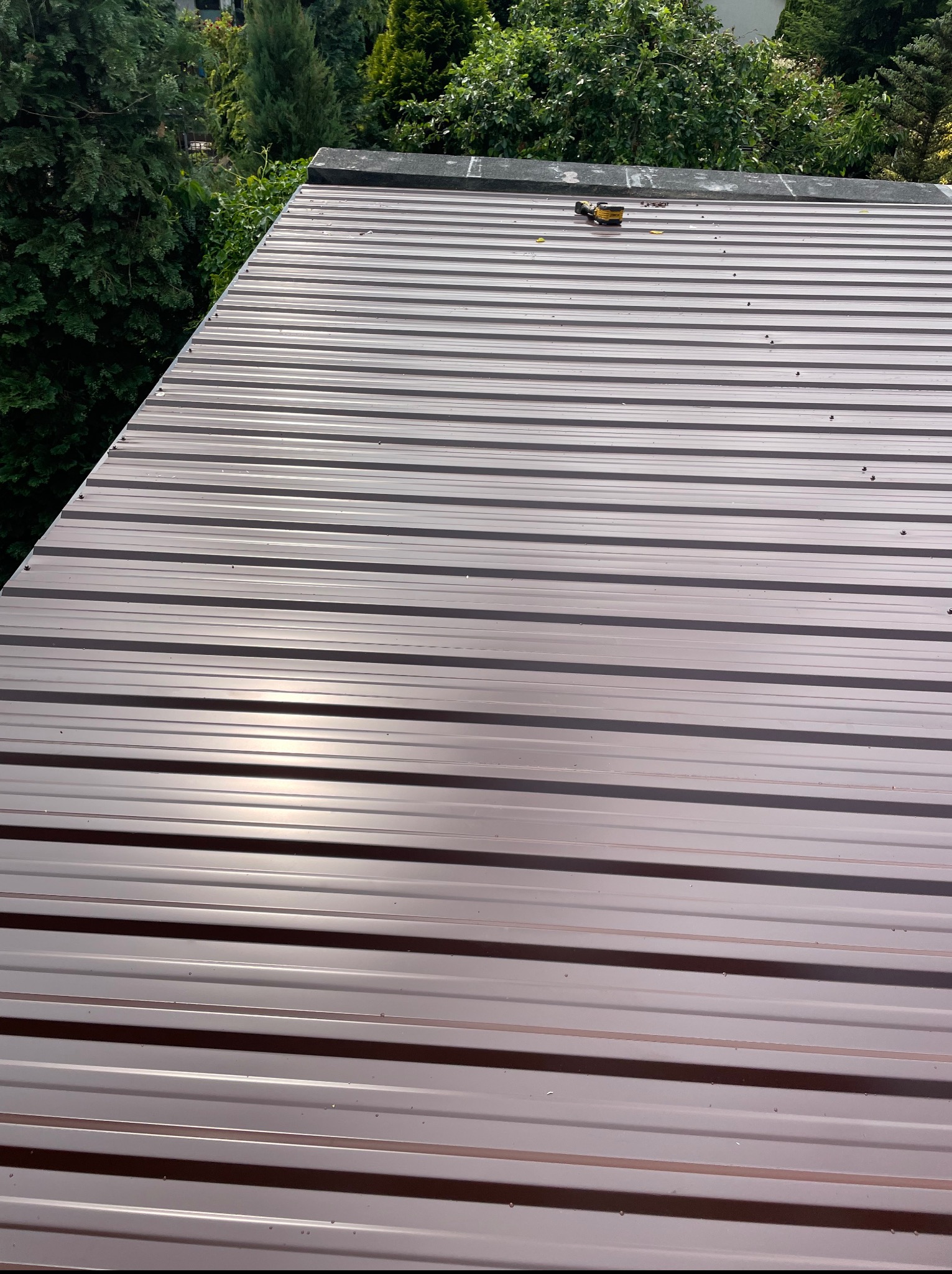 Błyszczący, brązowy dach z blachy trapezowej, widziany z góry pod kątem. Na dachu leży narzędzie. W tle zieleń drzew.