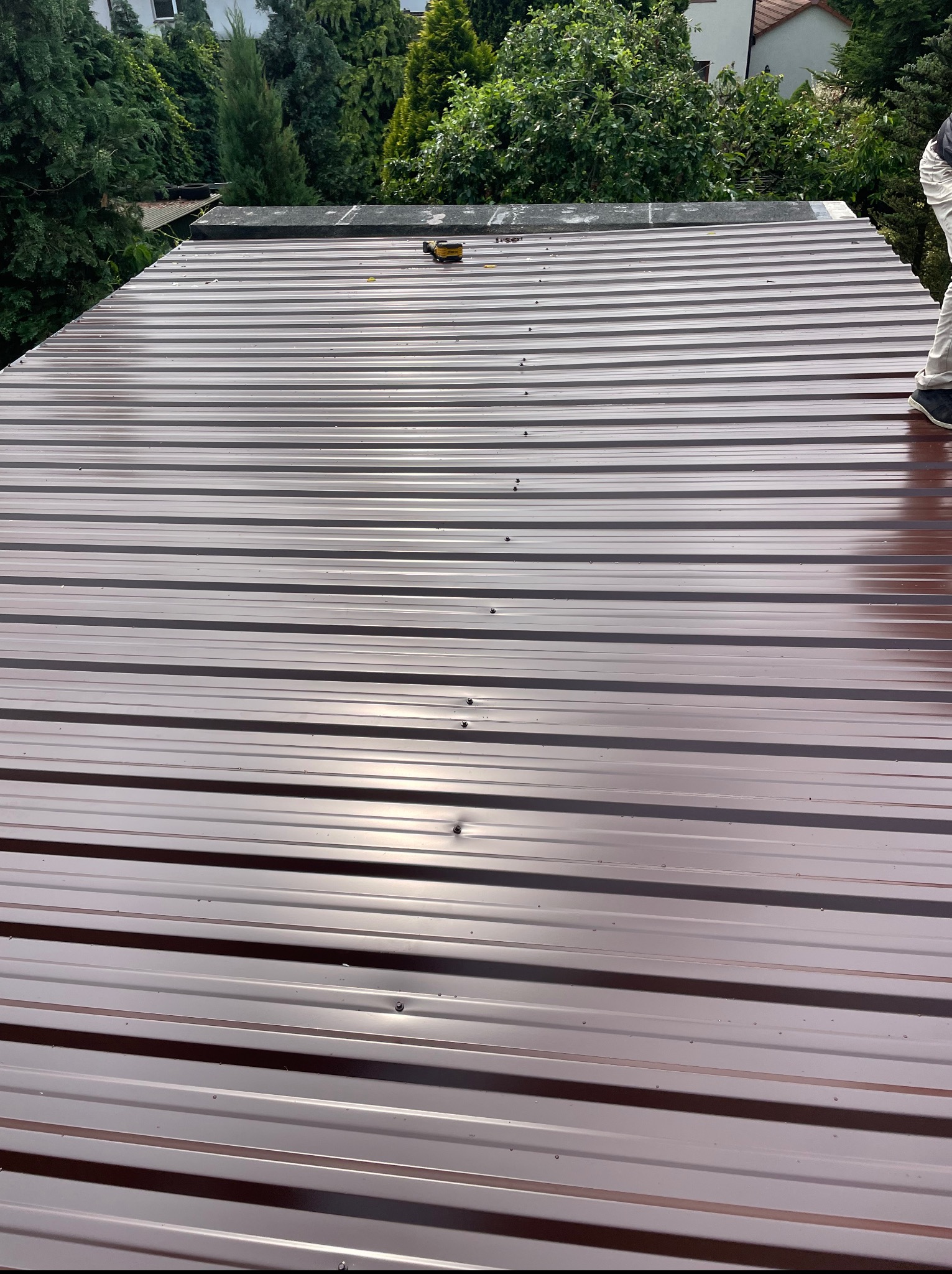 Błyszczący, brązowy dach z blachy trapezowej, z widocznymi śrubami mocującymi, częściowo mokry, z fragmentem osoby i narzędziem na tle zieleni.