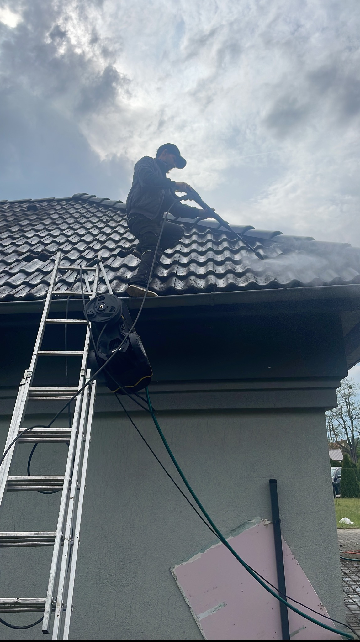 Pracownik z drabiny czyści dach z ciemnej dachówki za pomocą myjki ciśnieniowej. Widoczne zacieki i para wodna. Na ścianie ślady po naprawie elewacji.