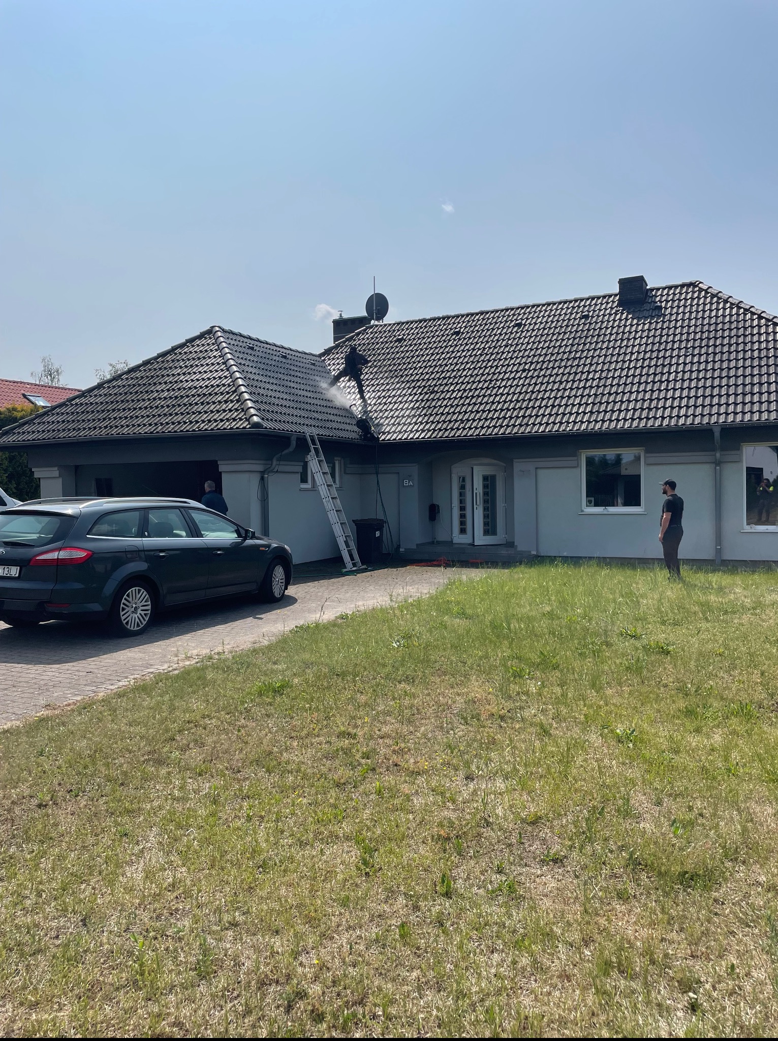 Dom jednorodzinny z szarą elewacją. Pracownik myje dach z drabiny, w tle zaparkowany samochód i trawnik. Słoneczny dzień.