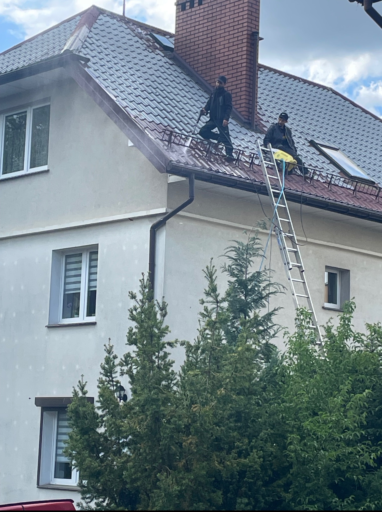 Dwóch mężczyzn czyści dach domu z metalową dachówką za pomocą myjki ciśnieniowej, stojąc na drabinie, z widocznym ceglanym kominem i oknami dachowymi.