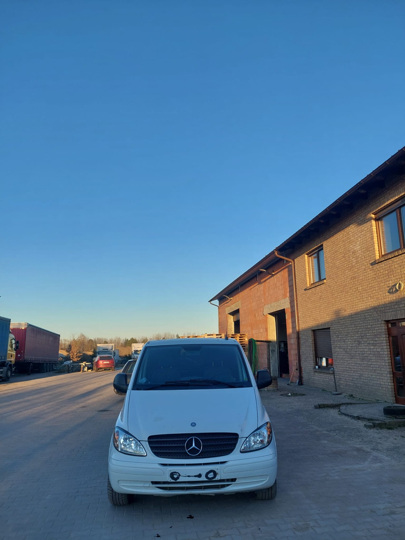 Biały Mercedes Vito na brukowanym placu, w tle budynek z cegły i inne pojazdy. Jasne niebo i popołudniowe światło.