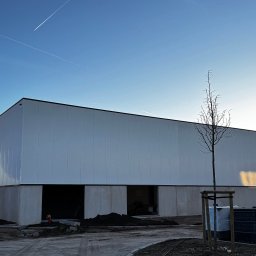 INTERICON CONSTRUCT SPÓŁKA Z OGRANICZONĄ ODPOWIEDZIALNOŚCIĄ - Nowoczesny budynek przemysłowy w trakcie budowy, z widocznymi otworami okiennymi i drzwiami, na tle błękitnego nieba z śladami smug kondensacyjnych samolotów.