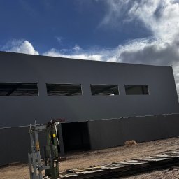 INTERICON CONSTRUCT SPÓŁKA Z OGRANICZONĄ ODPOWIEDZIALNOŚCIĄ - Szara hala w budowie z widocznymi oknami i sprzętem budowlanym na zewnątrz, pod błękitnym niebem z chmurami. Widok z dołu.