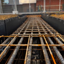 INTERICON CONSTRUCT SPÓŁKA Z OGRANICZONĄ ODPOWIEDZIALNOŚCIĄ - Z bliska: Zbrojenie fundamentów w wykopie z czarną folią, przygotowane do zalania betonem. Widok z dołu, na tle budynku i ogrodzenia budowy.