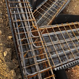 INTERICON CONSTRUCT SPÓŁKA Z OGRANICZONĄ ODPOWIEDZIALNOŚCIĄ - Zbrojenie fundamentów w szalunku z tworzywa, widok z góry. Stalowe pręty z widoczną rdzą, czarna folia wewnątrz szalunku, ziemia wokół. Etap prac fundamentowych.