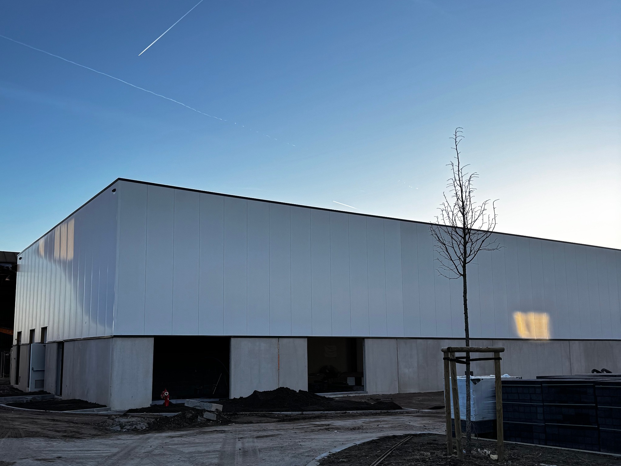 Nowoczesny budynek przemysłowy w trakcie budowy, z widocznymi otworami okiennymi i drzwiami, na tle błękitnego nieba z śladami smug kondensacyjnych samolotów.