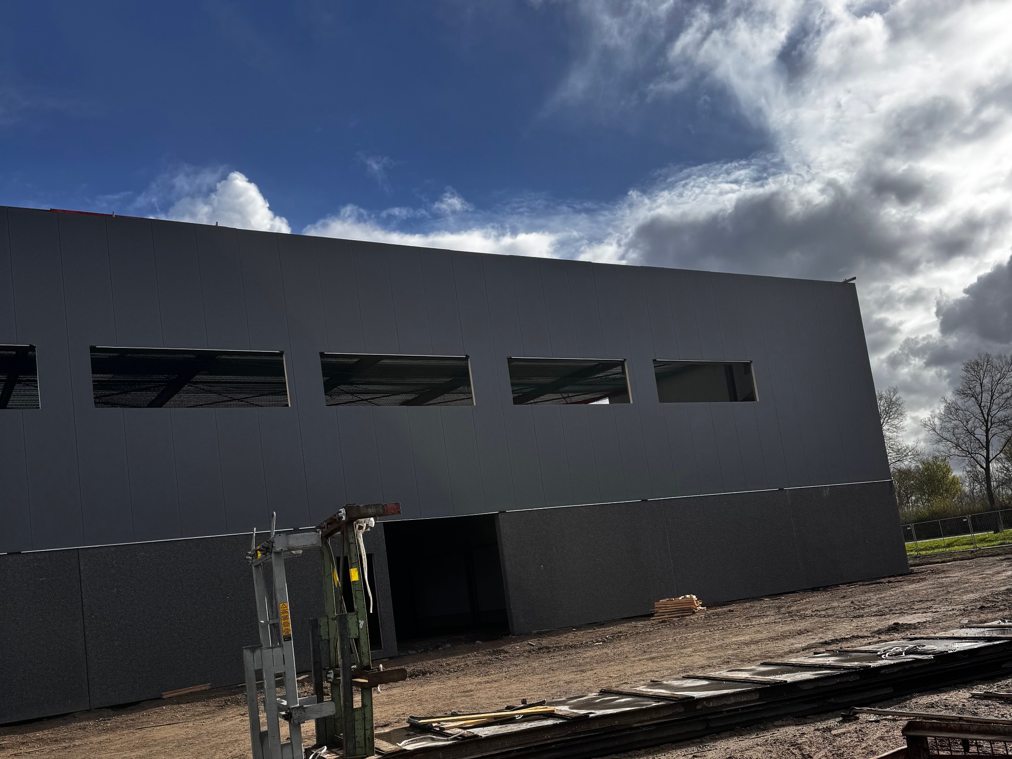 Szara hala w budowie z widocznymi oknami i sprzętem budowlanym na zewnątrz, pod błękitnym niebem z chmurami. Widok z dołu.