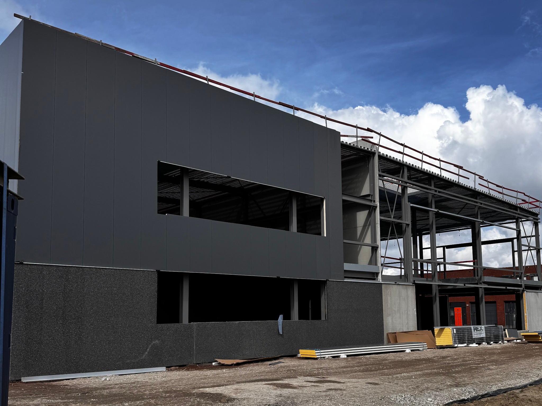 Budowa hali: stalowa konstrukcja, szare panele ścienne, widoczne okna i materiały budowlane. Błękitne niebo z chmurami w tle. Ujęcie z dołu, podkreślające skalę inwestycji.