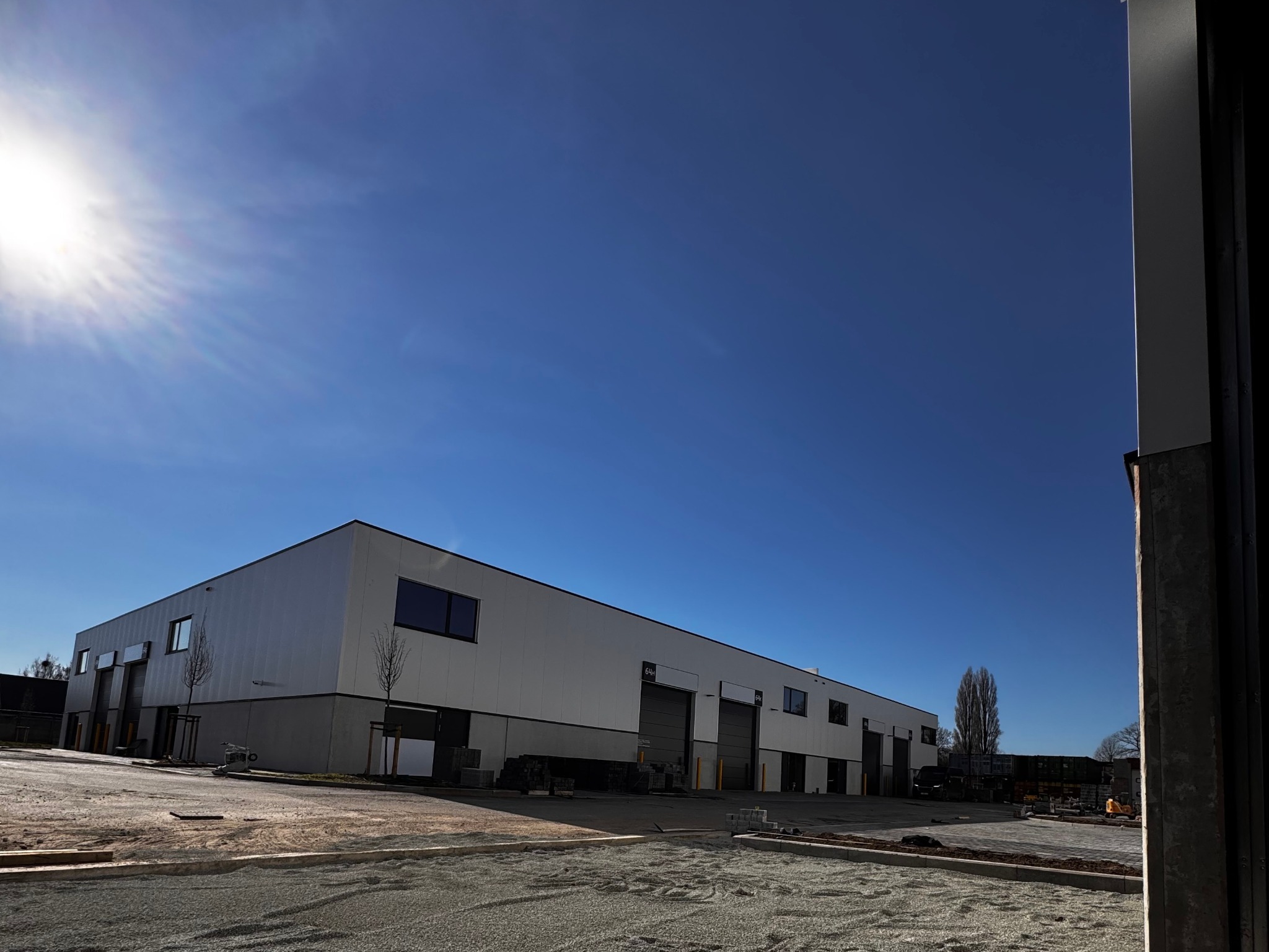 Nowoczesny budynek magazynowy z bramami rolowanymi w jasny dzień. Widoczne elementy instalacji zewnętrznej i plac manewrowy w trakcie budowy.