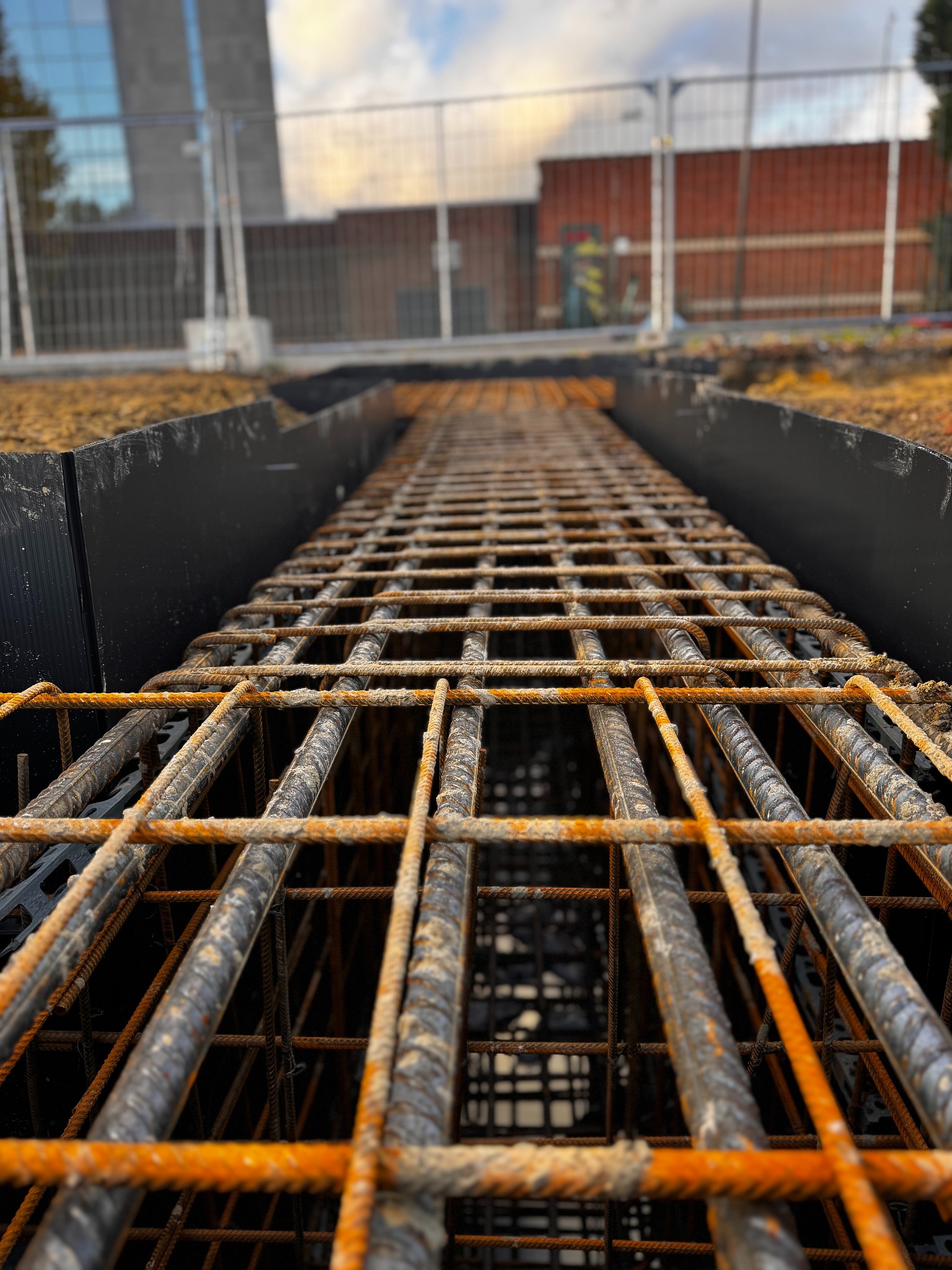 Z bliska: Zbrojenie fundamentów w wykopie z czarną folią, przygotowane do zalania betonem. Widok z dołu, na tle budynku i ogrodzenia budowy.