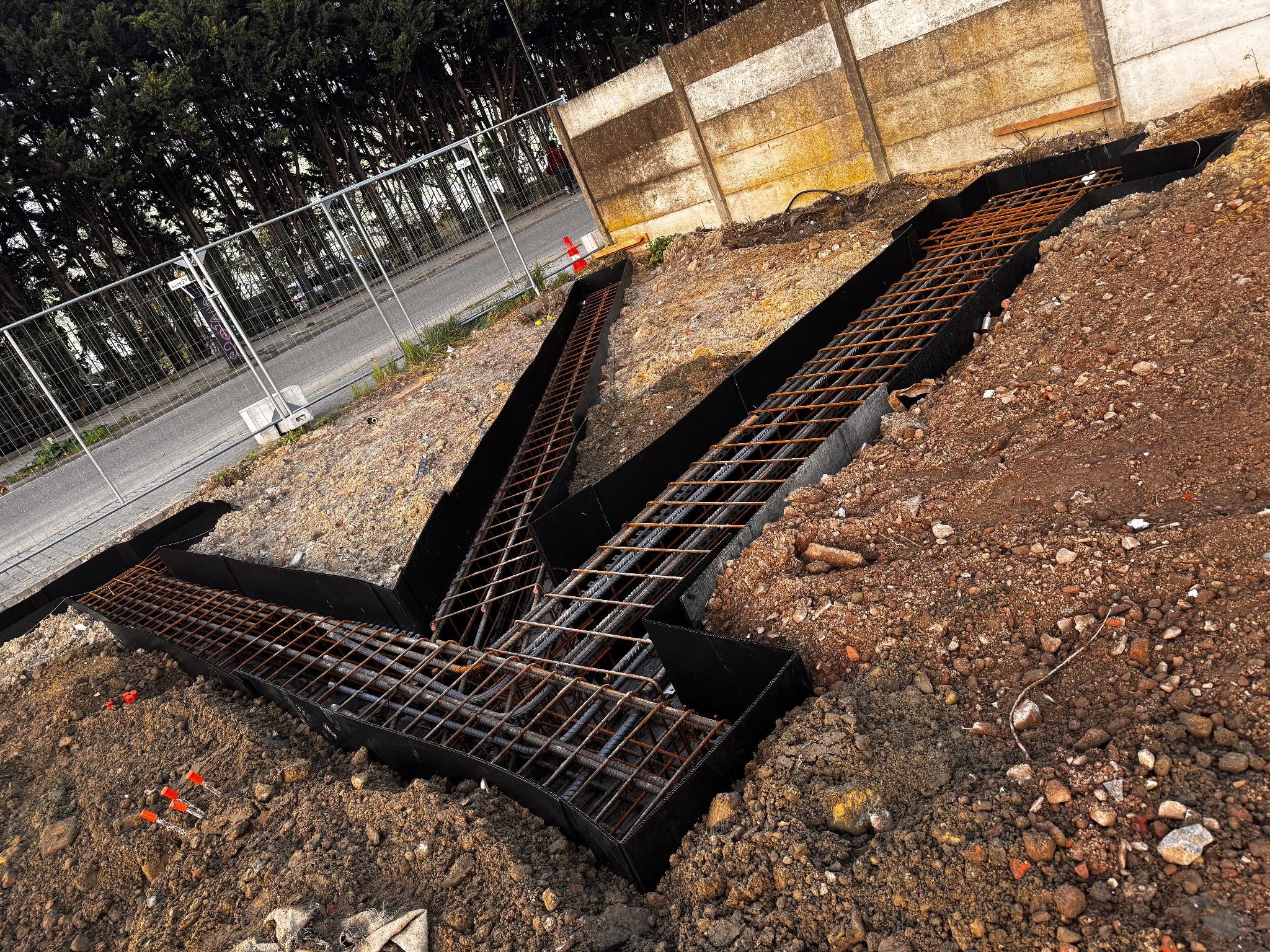 Zbrojenie fundamentów w wykopie, zabezpieczone czarną folią, przygotowane do zalania betonem. Widoczne elementy ogrodzenia i drzewa w tle.