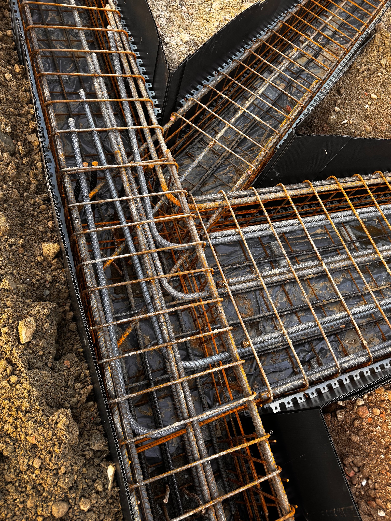 Zbrojenie fundamentów w szalunku z tworzywa, widok z góry. Stalowe pręty z widoczną rdzą, czarna folia wewnątrz szalunku, ziemia wokół. Etap prac fundamentowych.
