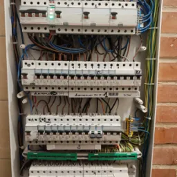 Rozdzielnica elektryczna wewnątrz obudowy, widoczne bezpieczniki, przewody, listwy zaciskowe i oznaczenia. Instalacja na tle ściany z cegły.