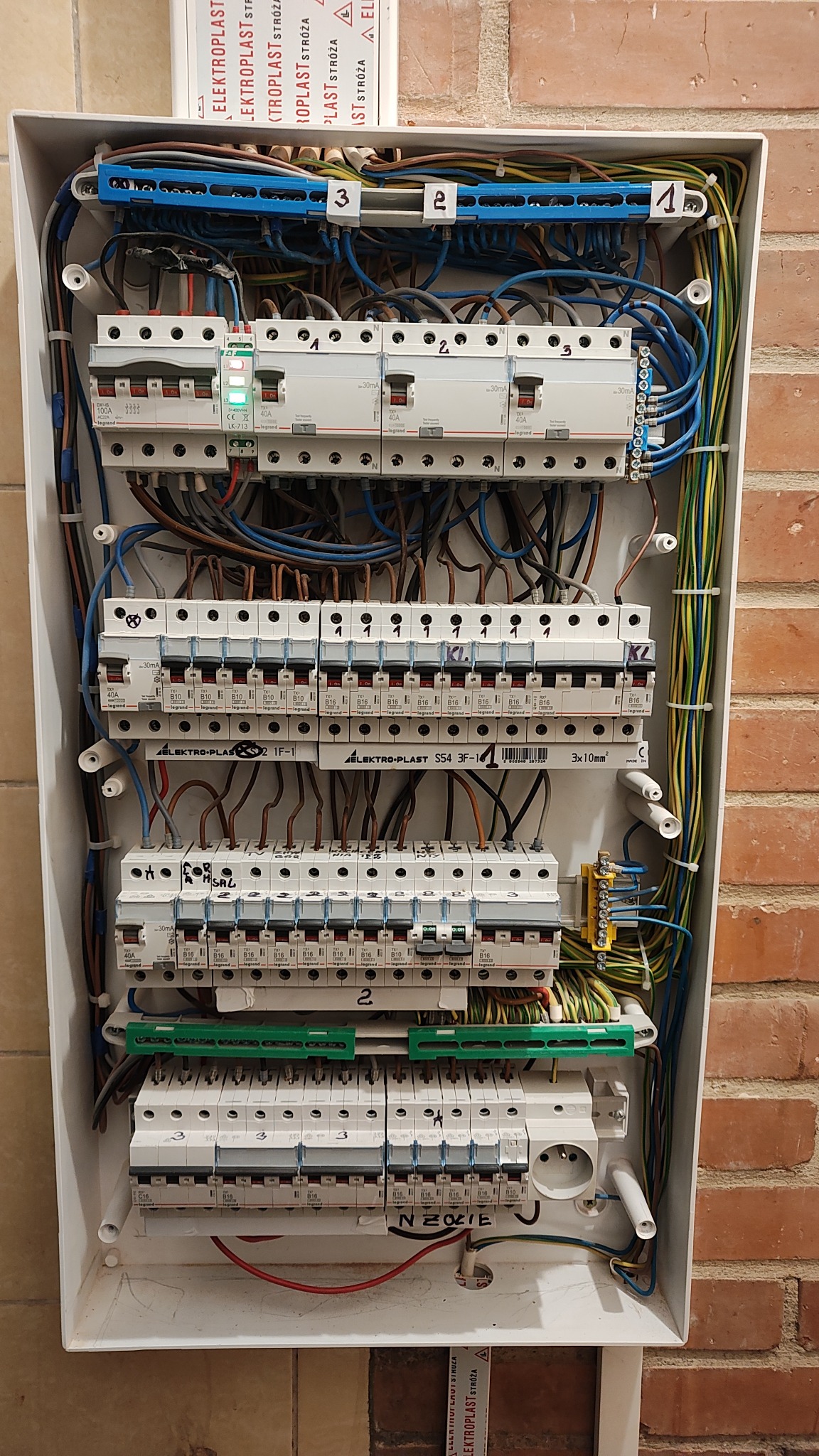 Rozdzielnica elektryczna wewnątrz obudowy, widoczne bezpieczniki, przewody, listwy zaciskowe i oznaczenia. Instalacja na tle ściany z cegły.