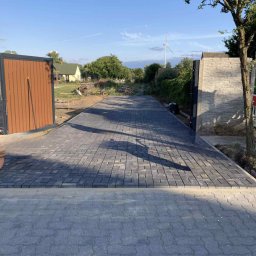 Frez-bud Paweł Jaszczuk - Ułożona szara kostka brukowa prowadząca do otwartej bramy wjazdowej, w tle widoczny dom i turbina wiatrowa. Słoneczny dzień.