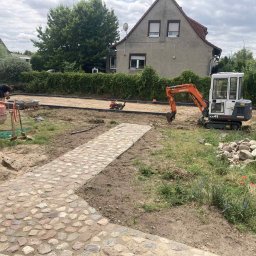Frez-bud Paweł Jaszczuk - Układanie kostki brukowej: mini koparka Kubota KX41, ubijarka, betonowa mieszarka i częściowo ułożony chodnik z kamienia polnego na posesji przed domem.
