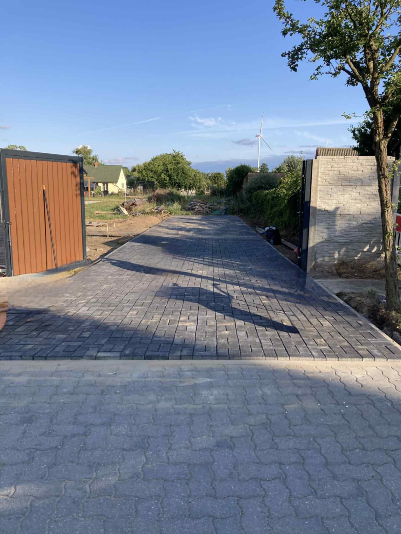 Ułożona szara kostka brukowa prowadząca do otwartej bramy wjazdowej, w tle widoczny dom i turbina wiatrowa. Słoneczny dzień.