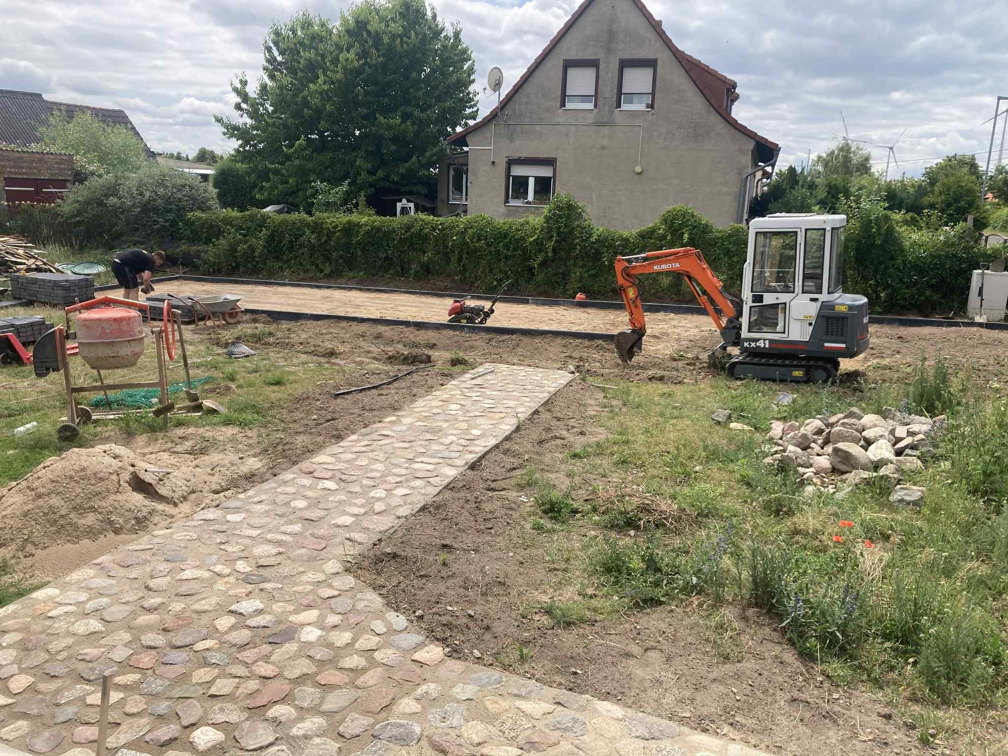 Układanie kostki brukowej: mini koparka Kubota KX41, ubijarka, betonowa mieszarka i częściowo ułożony chodnik z kamienia polnego na posesji przed domem.
