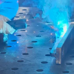 Praca spawacza w trakcie łączenia elementów metalowych na perforowanym stole spawalniczym, w tle ochronna maska i kłęby dymu.