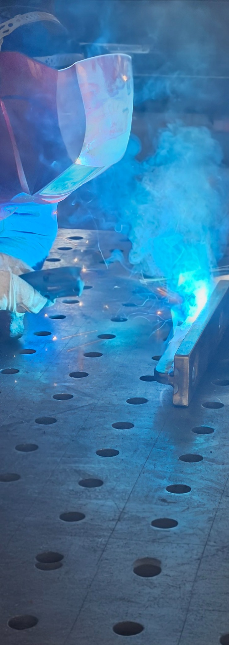 Praca spawacza w trakcie łączenia elementów metalowych na perforowanym stole spawalniczym, w tle ochronna maska i kłęby dymu.