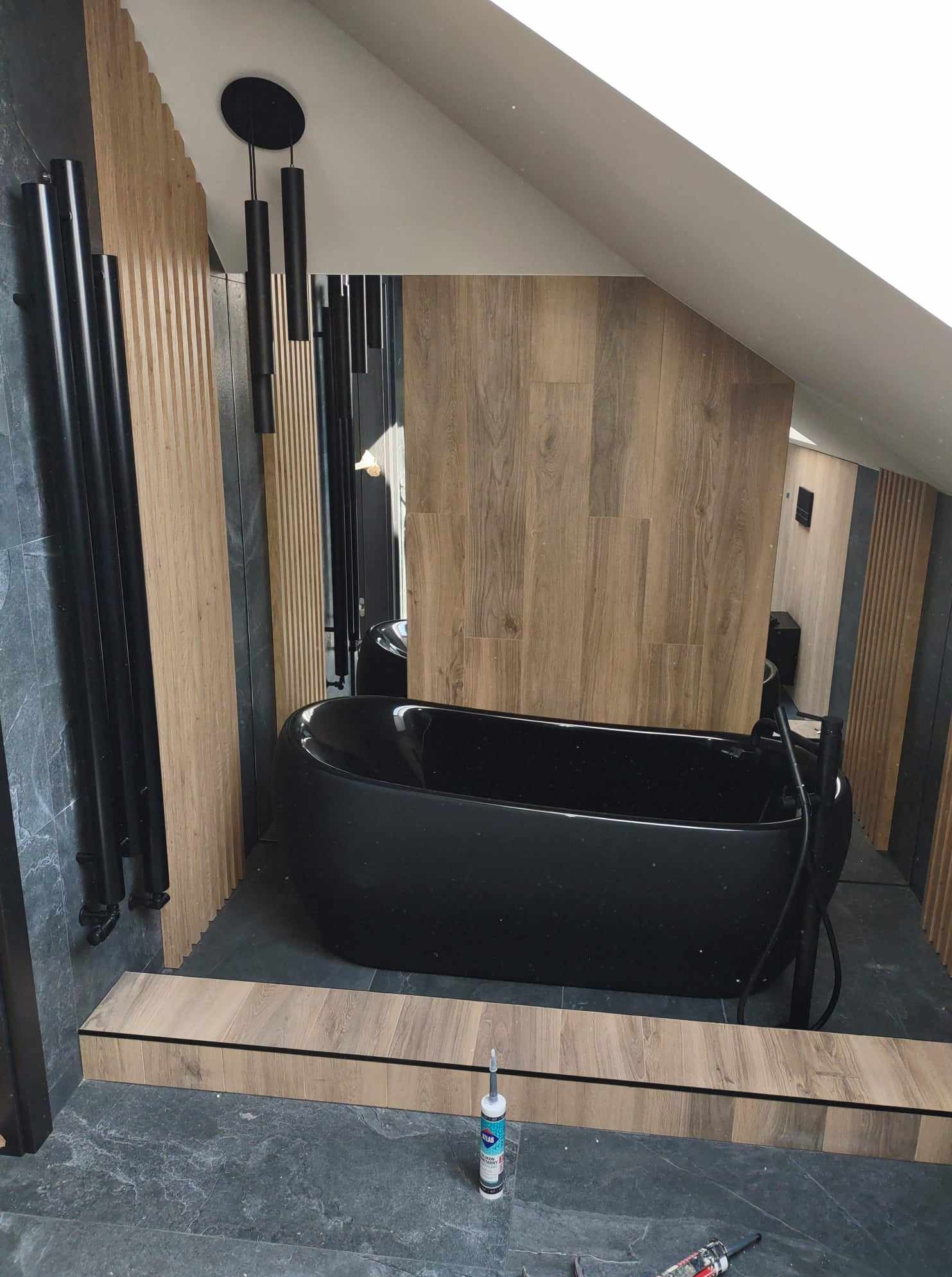 Nowoczesna łazienka na poddaszu z czarną wanną wolnostojącą, drewnianymi lamelami i grafitową płytką. Widoczny silikon i narzędzia, co sugeruje trwające prace wykończeniowe.