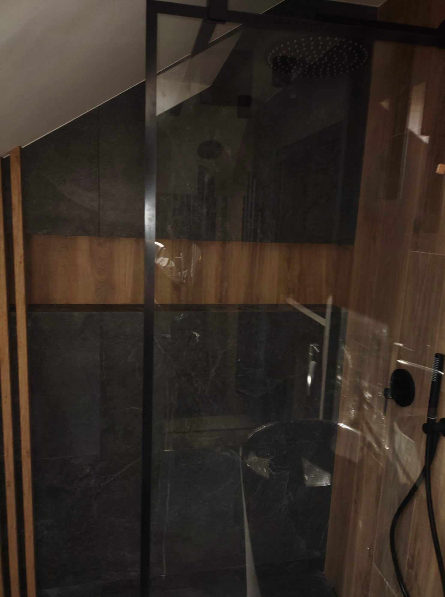 Nowoczesna kabina prysznicowa z ciemnymi płytkami imitującymi marmur i drewnianymi akcentami, widoczna armatura w czarnym macie.
