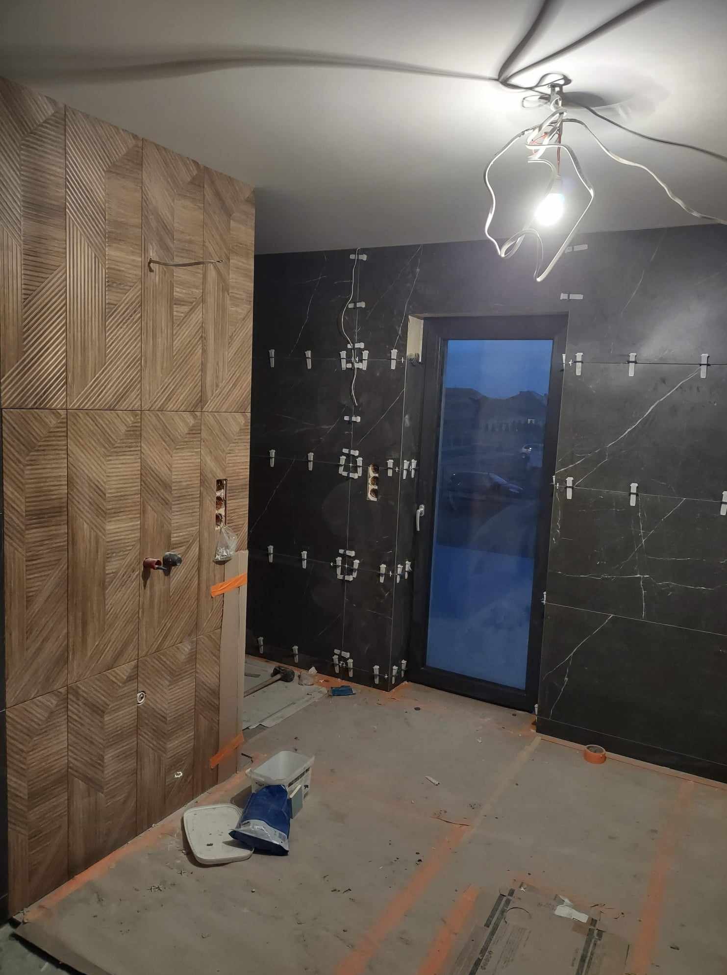 Wnętrze łazienki w trakcie prac: ściany wyłożone ciemnymi płytkami z marmurowym wzorem i drewnopodobnymi płytkami ułożonymi we wzór jodełki. Widoczne narzędzia i elementy montażowe.