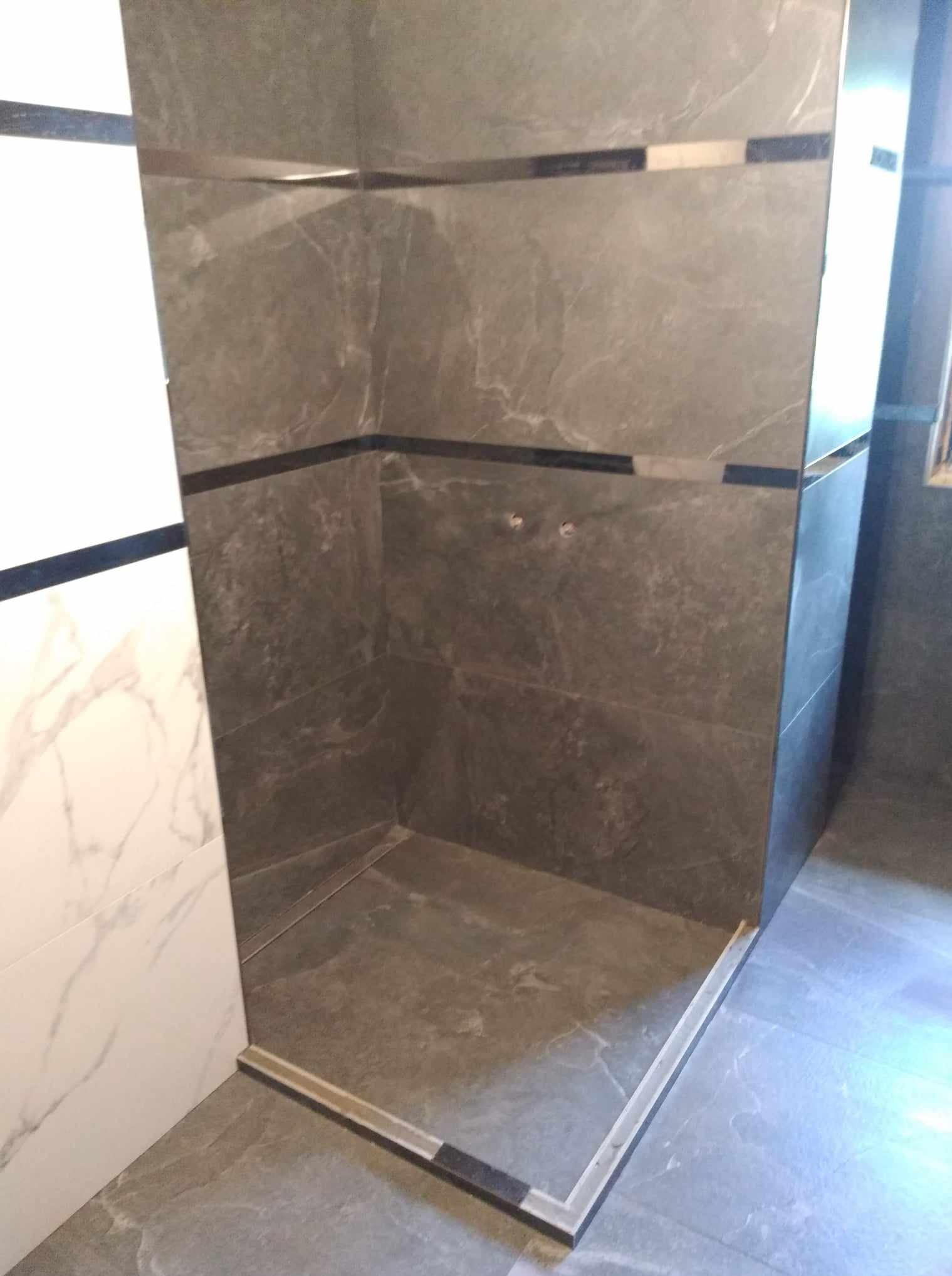 Nowoczesny prysznic walk-in z szarymi płytkami imitującymi kamień i białymi płytkami marmurkowymi. Widoczne listwy dekoracyjne i odpływ liniowy.