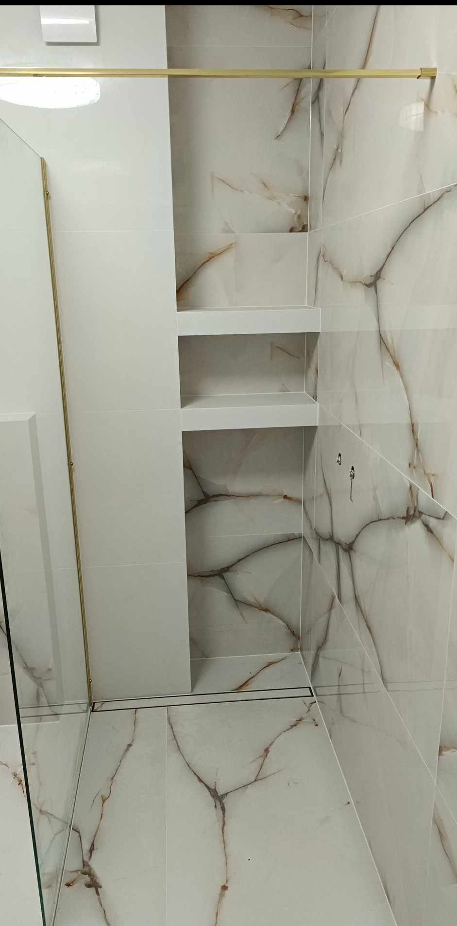 Wykończona łazienka z marmurowymi płytkami, złotymi akcentami i wnęką z półkami. Nowoczesny design, minimalistyczny styl, widoczny odpływ liniowy.
