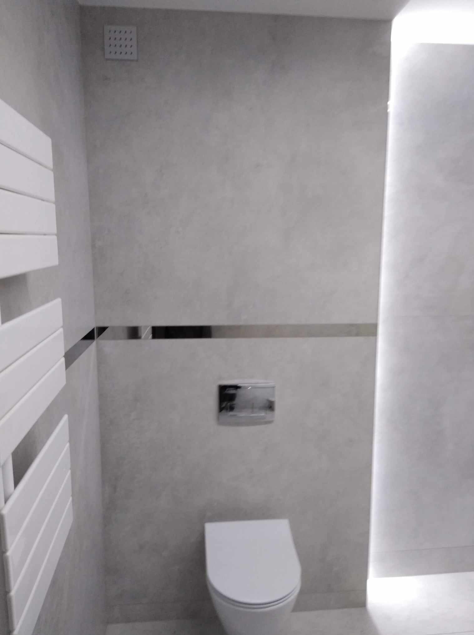 Wykończona łazienka w szarościach: płytki imitujące beton, podświetlenie LED wzdłuż krawędzi, chromowana listwa dekoracyjna i grzejnik drabinkowy.