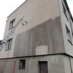 Mycie elewacji w Lublinie. Fragment umytej, szarej ściany budynku z fakturą, z drabiny z dyszami. Widoczna dolna część budynku z drzwiami i oknem.