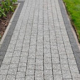 Kubuś - Szara kostka brukowa ułożona wzdłuż ścieżki, obrzeża z ciemniejszej kostki, trawnik po prawej, budynek z czerwoną cegłą po lewej stronie. Ujęcie z perspektywy.