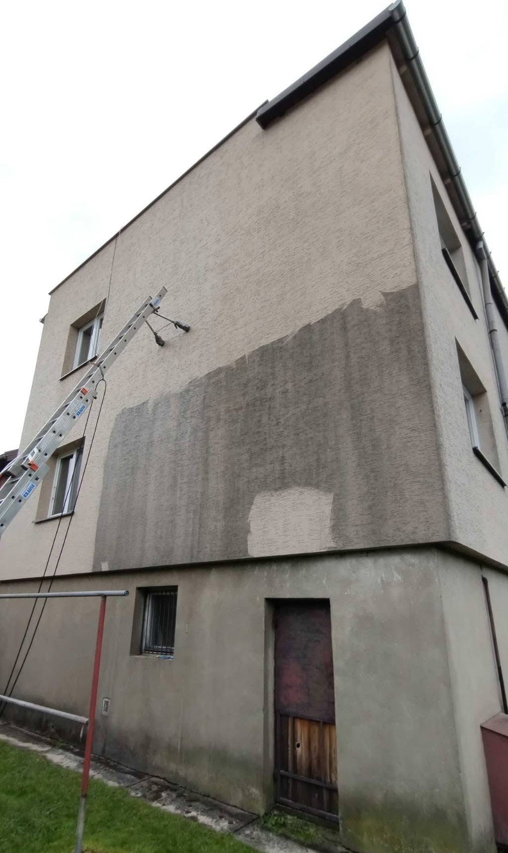 Mycie elewacji w Lublinie. Fragment umytej, szarej ściany budynku z fakturą, z drabiny z dyszami. Widoczna dolna część budynku z drzwiami i oknem.