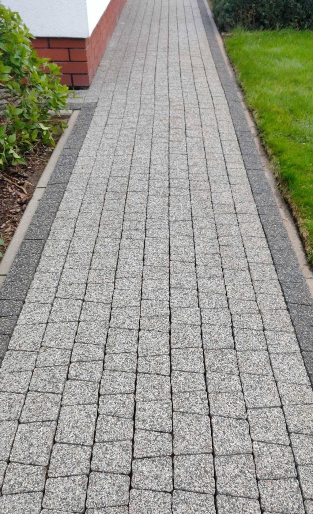 Szara kostka brukowa ułożona wzdłuż ścieżki, obrzeża z ciemniejszej kostki, trawnik po prawej, budynek z czerwoną cegłą po lewej stronie. Ujęcie z perspektywy.