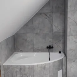 Wykończona łazienka na poddaszu ze skośnym sufitem. Szare płytki imitujące marmur na ścianach i podłodze, biała narożna wanna z czarną armaturą.