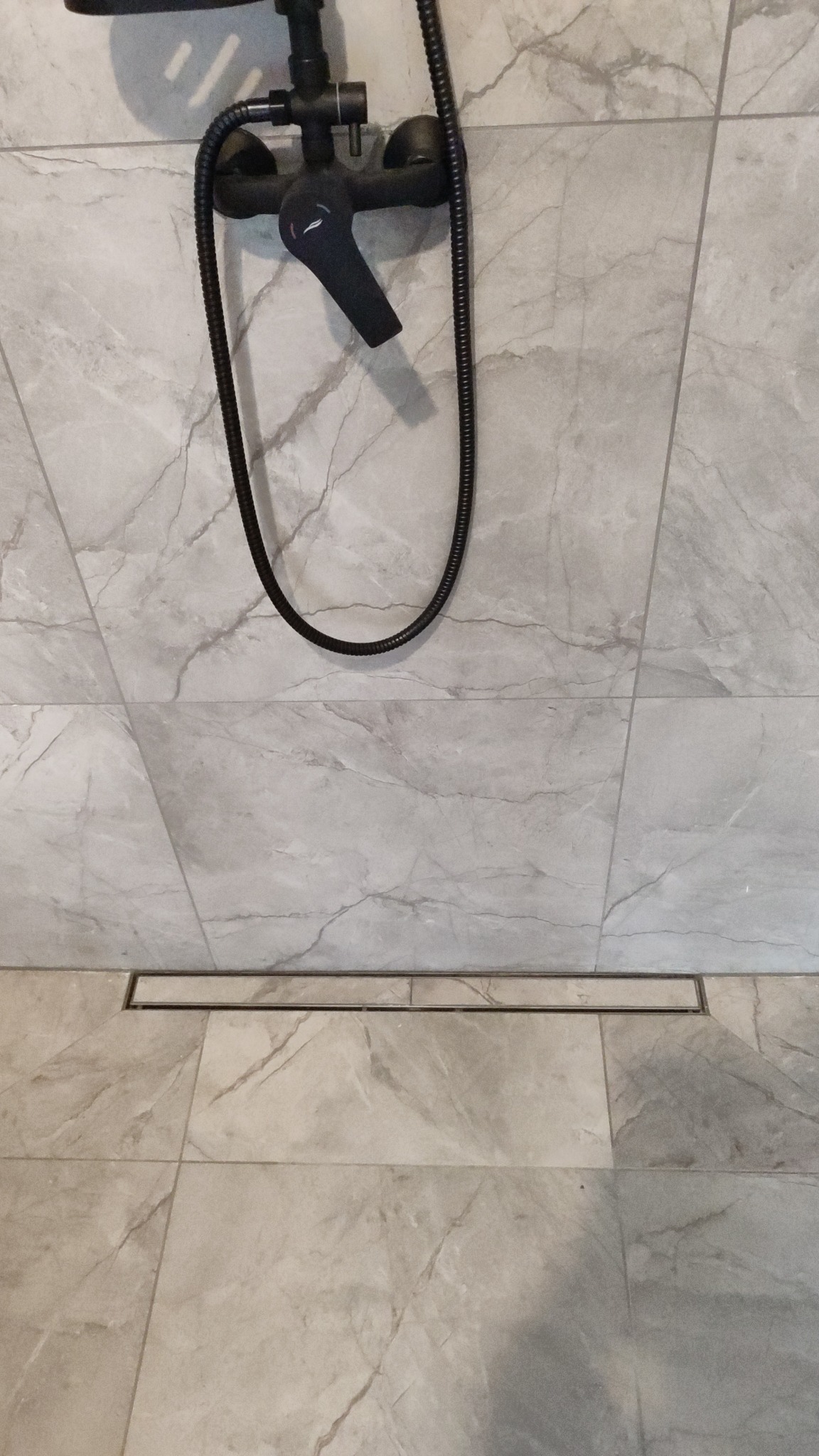 Szara łazienka z marmurowymi płytkami, czarną armaturą i odpływem liniowym. Minimalistyczny design, eleganckie wykończenie. Widok na dół ściany i podłogę.