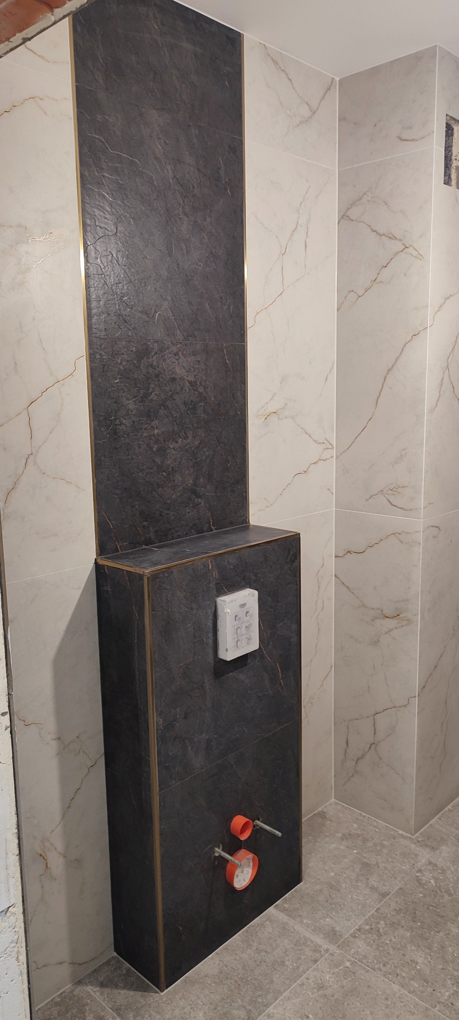 Wykończona łazienka: zabudowa stelaża WC z czarnej płyty imitującej kamień, obok jasne płytki z marmurowym wzorem, złote listwy. Widoczne przyłącza hydrauliczne.