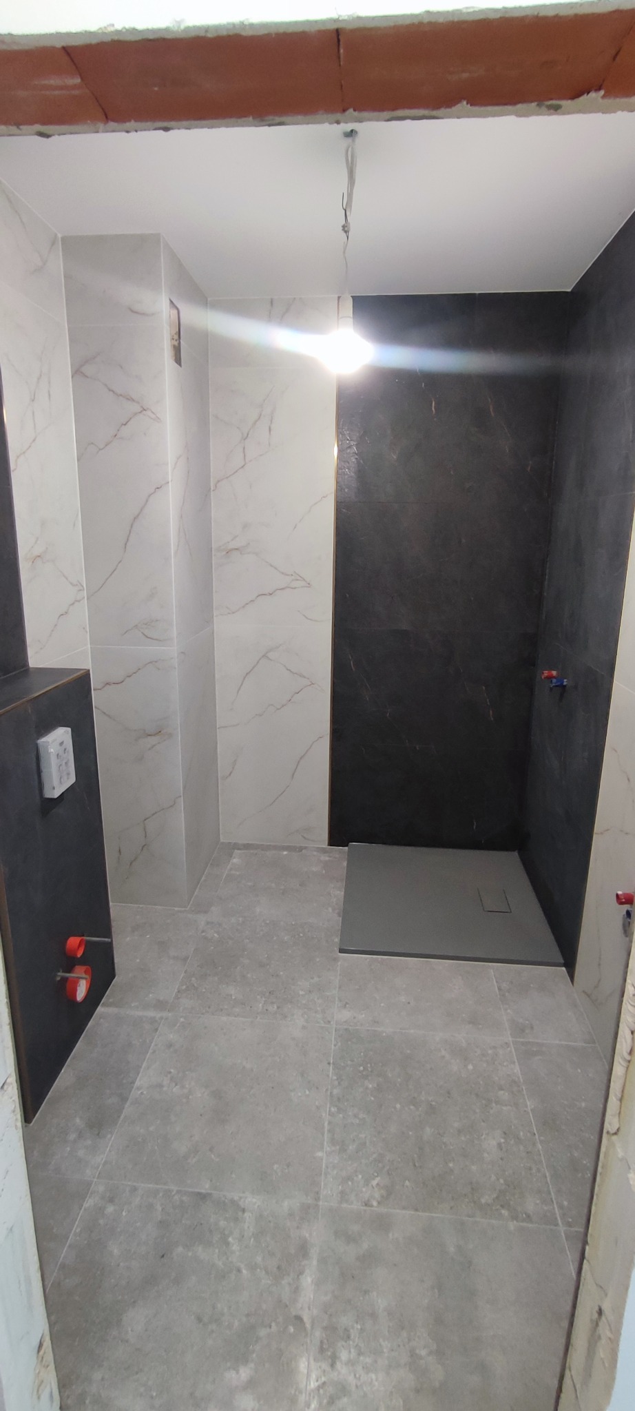 Wykończona łazienka z szarymi płytkami podłogowymi i marmurowymi płytkami ściennymi, kabina prysznicowa z czarną ścianą, widoczne instalacje wodne.