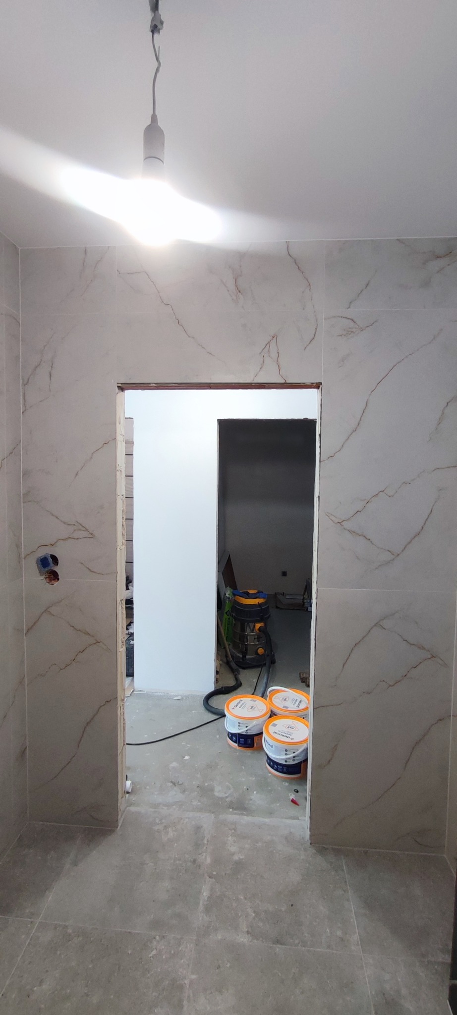 Wykończona łazienka z marmurowymi płytkami, widok na sąsiednie pomieszczenie z odkurzaczem i farbami. Surowa żarówka zwisa z sufitu.