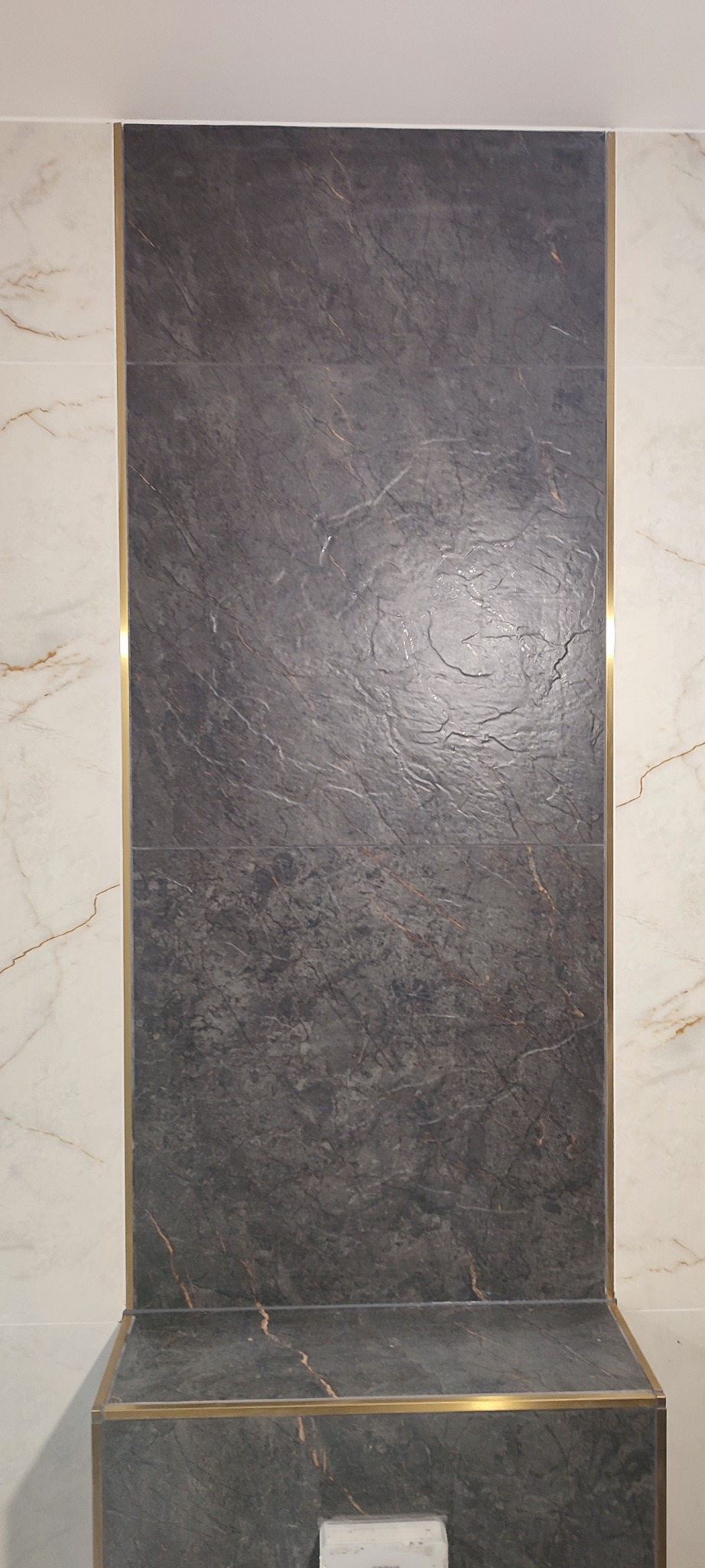 Pionowy fragment ściany z szarej płytki imitującej kamień, obramowanej złotą listwą. Po bokach widoczne jasne płytki z marmurkowym wzorem. Detal wykończenia łazienki.